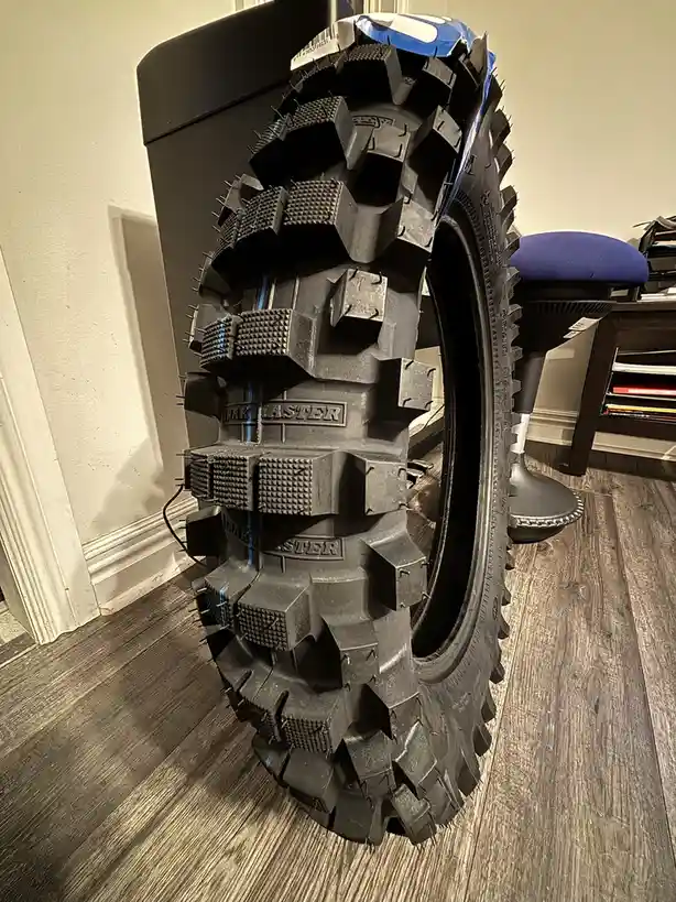 A large, rugged, black off-road tire with deep treads and spikes is standing upright on a wooden floor indoors, next to a…