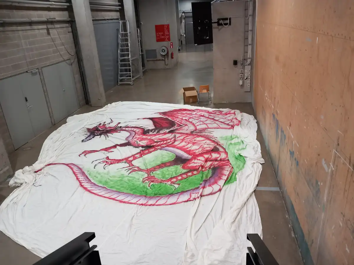 A large white fabric is spread across a concrete floor in an industrial-looking hallway, featuring a detailed red dragon…