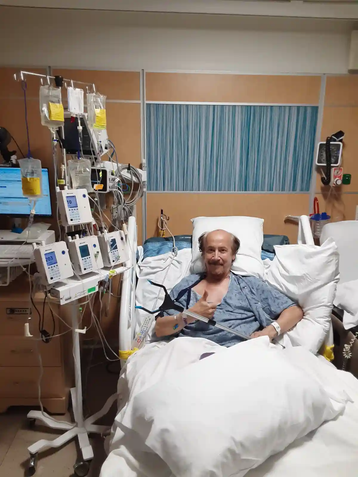 An older man with a mustache lies in a hospital bed surrounded by medical equipment, including IV bags, monitors, and a…