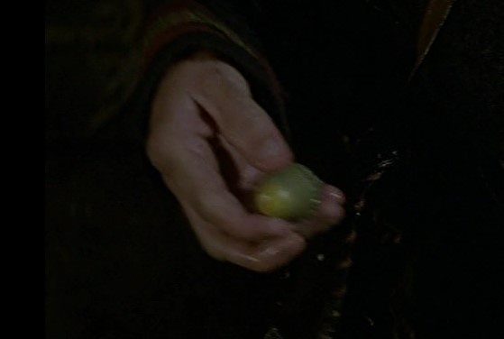 A dimly lit close-up shows a person's hand holding a small, greenish-yellow fruit, possibly a grape or small plum, against a…