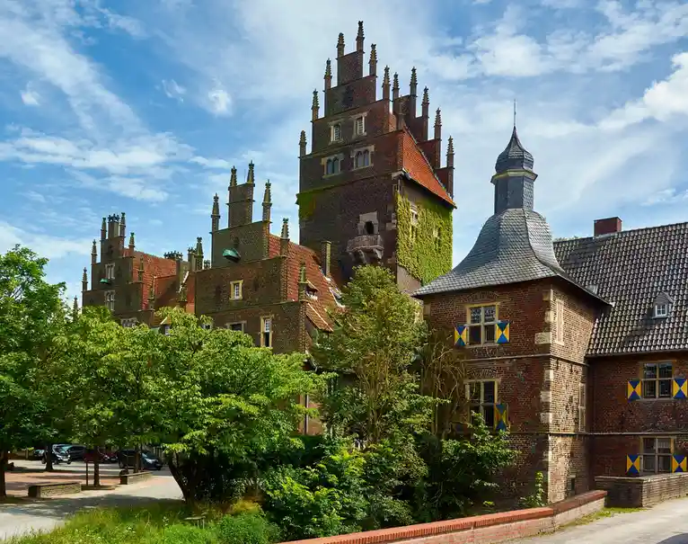 A picturesque photo of a large, ornate brick castle with red-tiled roofs, ivy-covered walls, and multiple turrets under a…