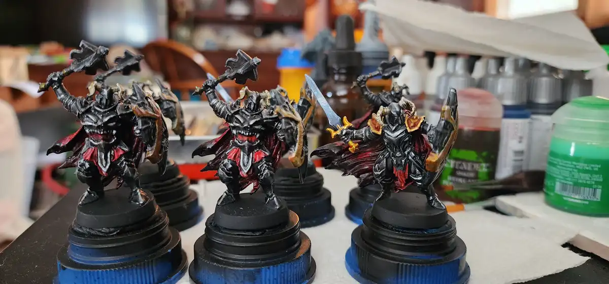 Three identical fantasy miniatures of demonic warriors, each holding a weapon and wearing black armor with red and gold…