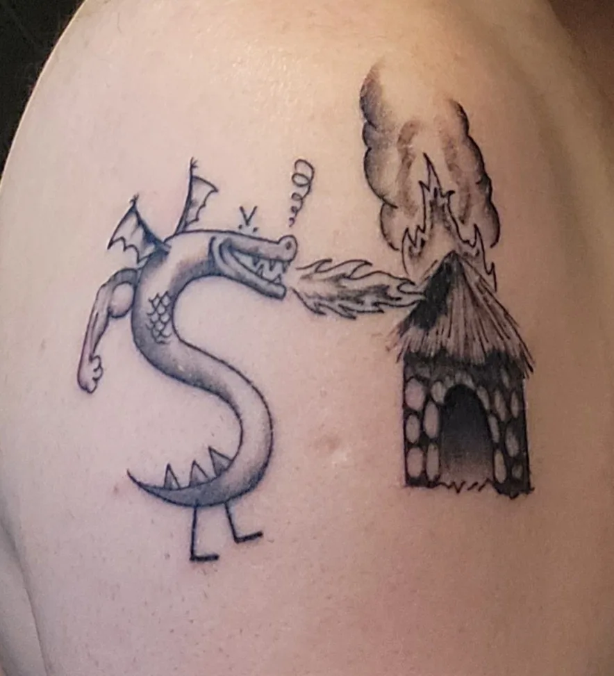 A black and grey tattoo on human skin depicts a cartoonish dragon whose body is shaped like a dollar sign, breathing fire…