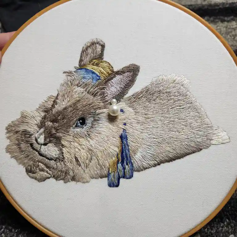 A detailed embroidery of a realistic-looking brown rabbit on white fabric held in a wooden hoop, adorned with a pearl…