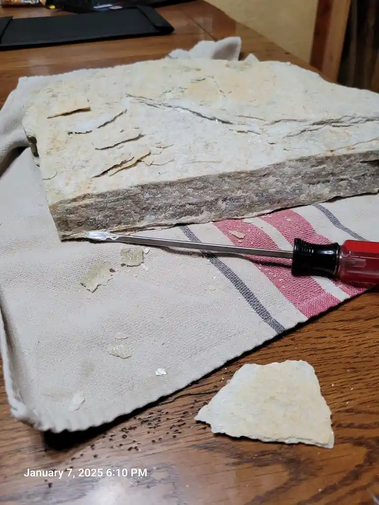A large, layered, flaky white rock sits on a striped cloth on a wooden table, with a screwdriver resting beside it and small…