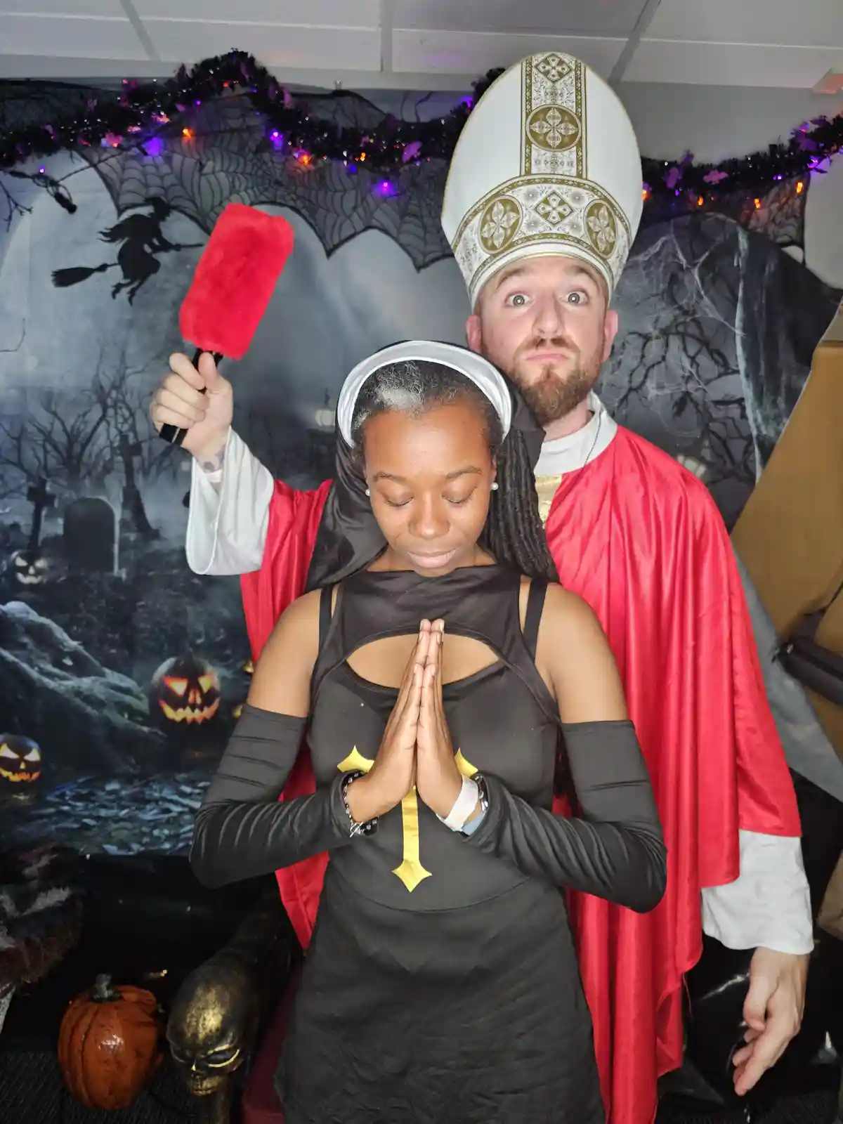 A man dressed as a Catholic priest with a red cape and mitre stands behind a woman in a black nun costume with her eyes…