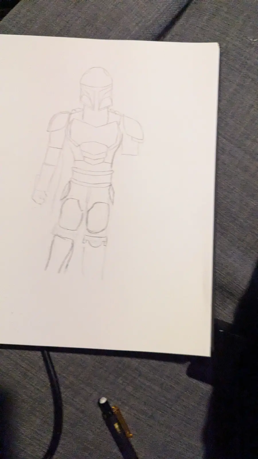 A pencil sketch on white paper depicts a figure in Mandalorian armor, viewed from the front. The drawing is a rough outline…