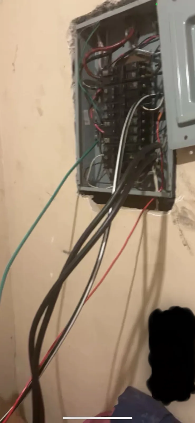 A close-up photo shows an open electrical breaker box mounted on a beige wall, with numerous red, black, green, and white…