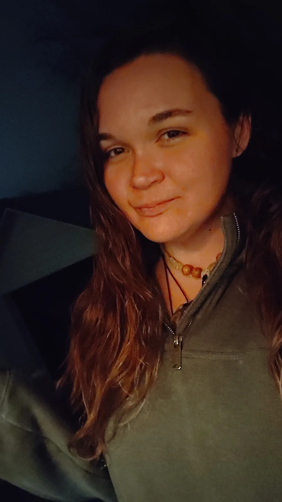 A close-up selfie of a woman with long brown hair, wearing a green zip-up hoodie and a beaded necklace, smiling slightly in…