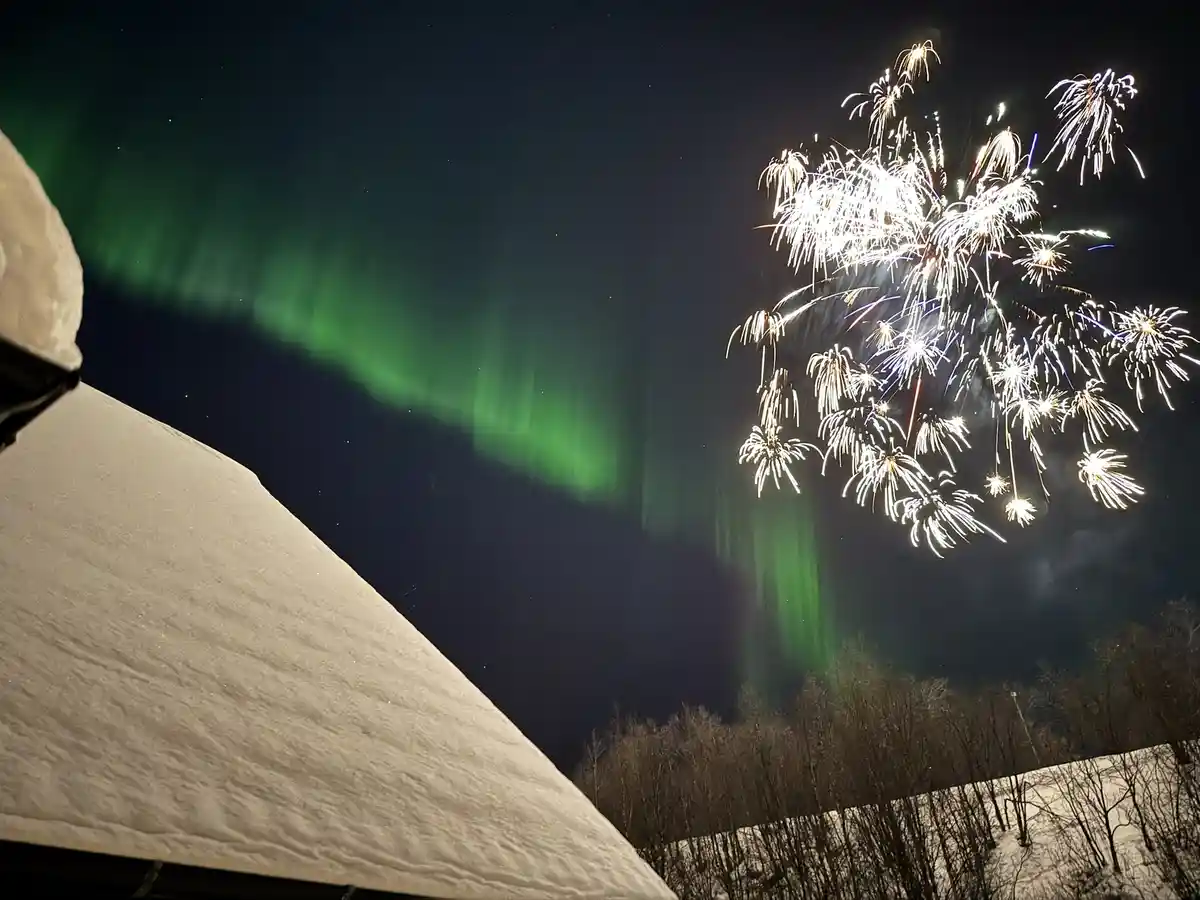 A snowy landscape at night shows a vibrant green aurora borealis arching across the sky, partially obscured by a large burst…