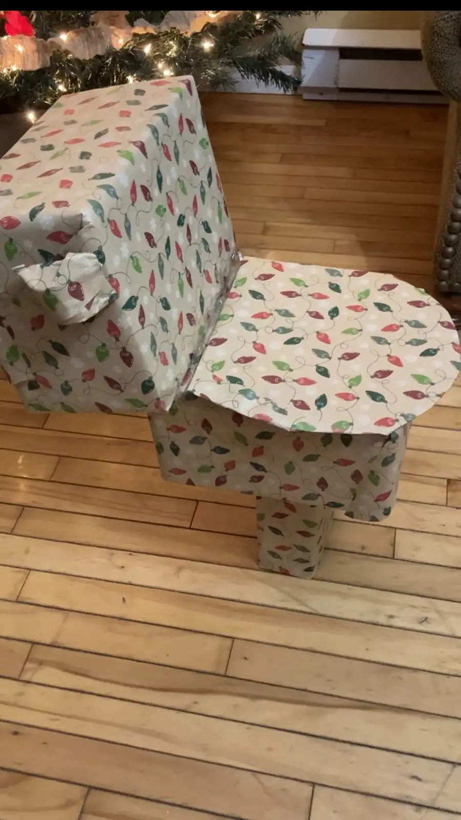 A piano is entirely wrapped in festive Christmas wrapping paper featuring red, green, and white light bulb patterns, with a…