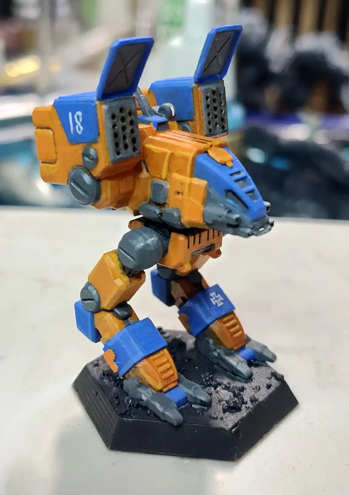 A painted orange and blue miniature robot model stands on a black hexagonal base, its back-mounted weapon systems open and…