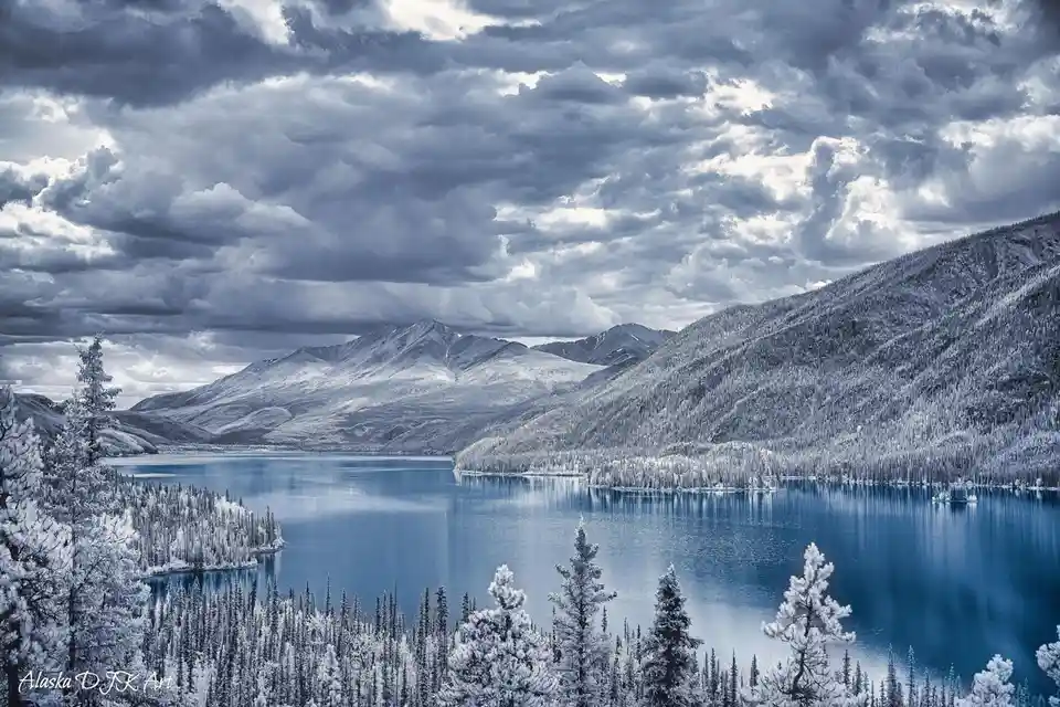 A serene, monochromatic winter landscape photograph of a snow-covered mountain range and a calm blue lake, with frosted…