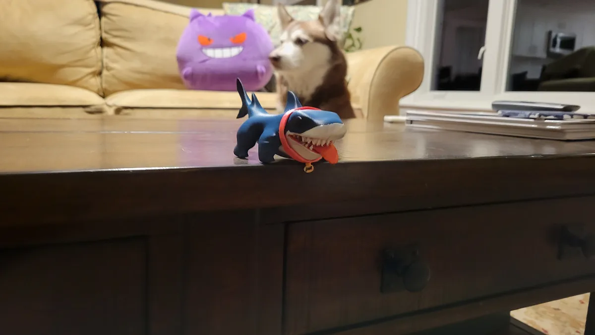 A blue plastic shark toy with an open mouth and red collar sits on a wooden coffee table. In the background, a husky dog…