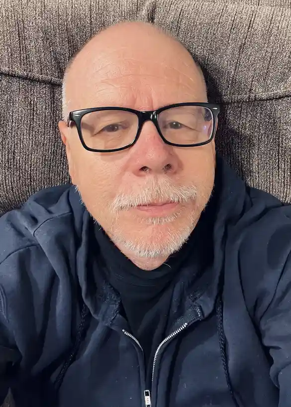 A close-up selfie of a middle-aged to older man with a balding head, wearing black-rimmed glasses and a dark blue zip-up…
