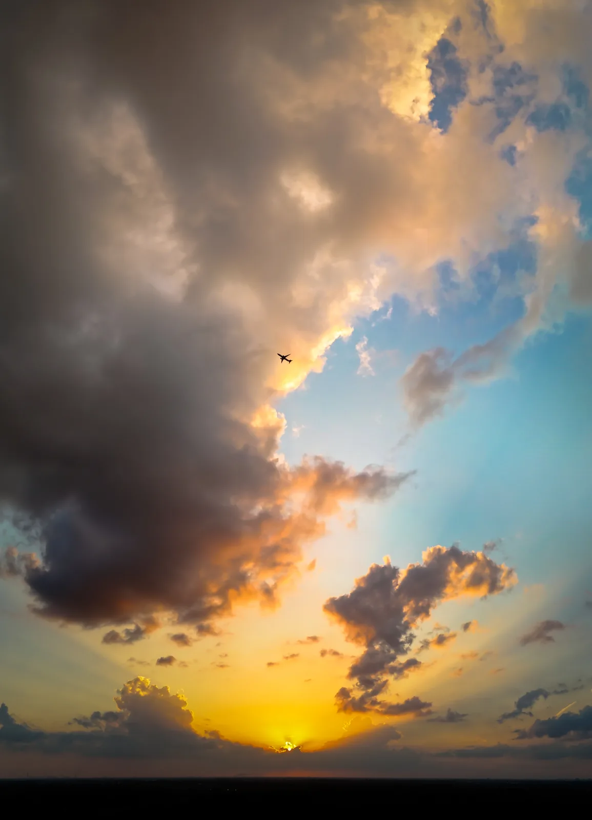 A silhouette of an airplane is visible against a dramatic sunset sky filled with orange, yellow, and blue clouds. The sun is…