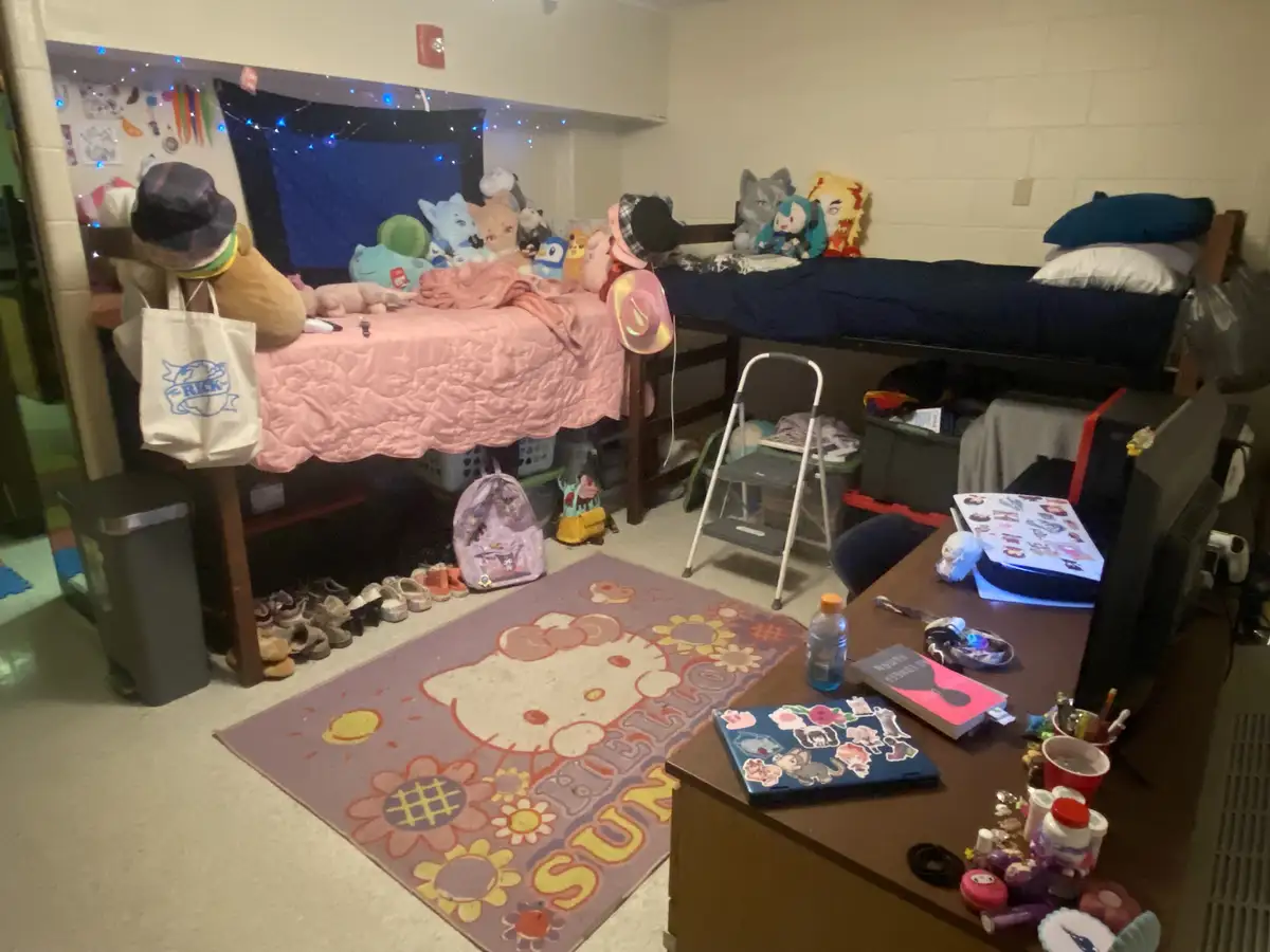 A messy college dorm room with two beds, one covered in pink bedding and stuffed animals, the other in blue. A Hello Kitty…