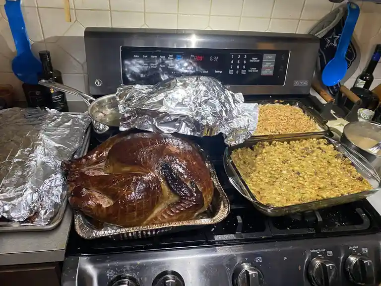 A roasted turkey sits on a stovetop next to two glass dishes of stuffing, all covered in foil or on trays, with an oven…