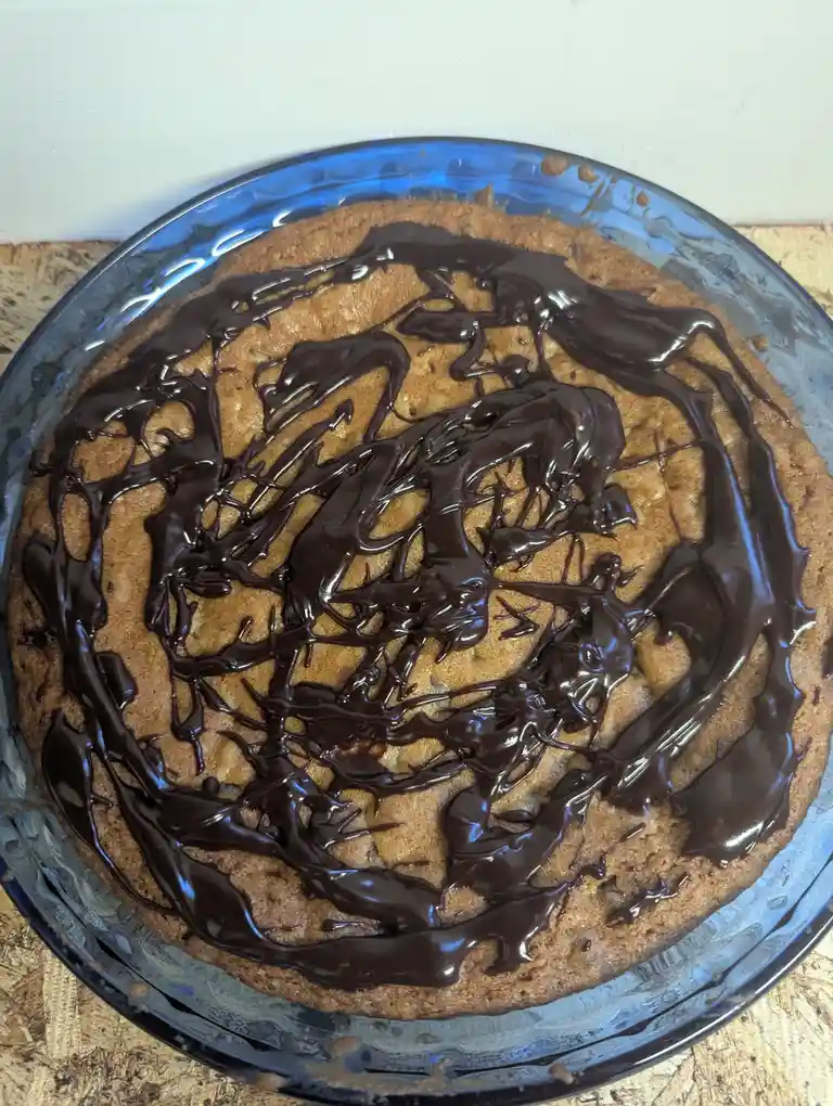 A round baked cake in a blue glass dish is covered in a messy, drizzled pattern of melted chocolate, resembling a chaotic…