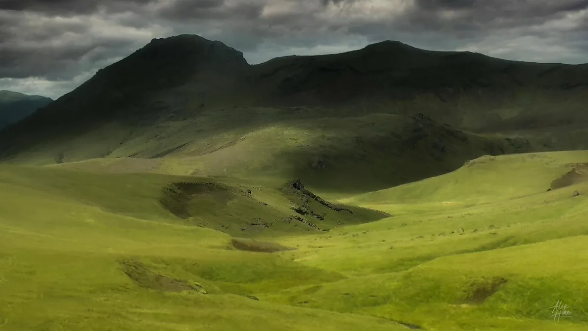 A scenic landscape photo of rolling green hills under a dark, cloudy sky, with a watermark in the bottom right corner…