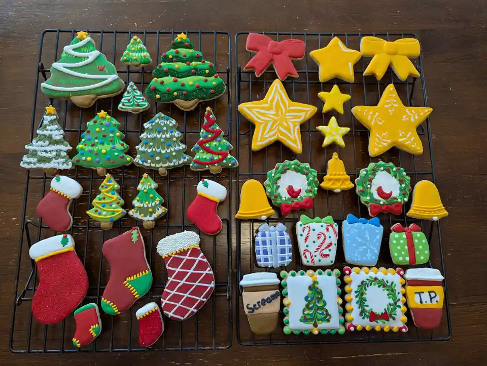 Two black wire cooling racks hold dozens of elaborately decorated Christmas-themed sugar cookies on a wooden table. The left…