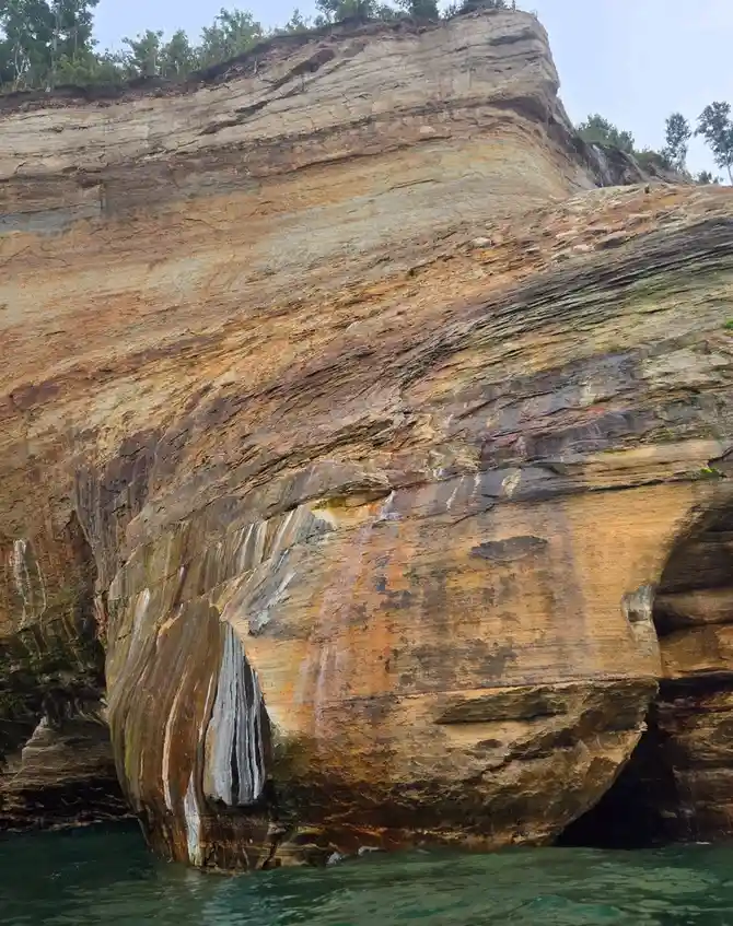 a natural rock formation by the water has distinct layers and shapes that resemble a human face, with what looks like eyes…