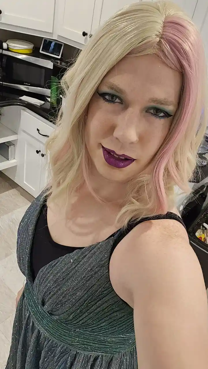 A person with blonde hair streaked pink, wearing bold teal eyeshadow and glittery purple lipstick, poses in a sparkly dark…
