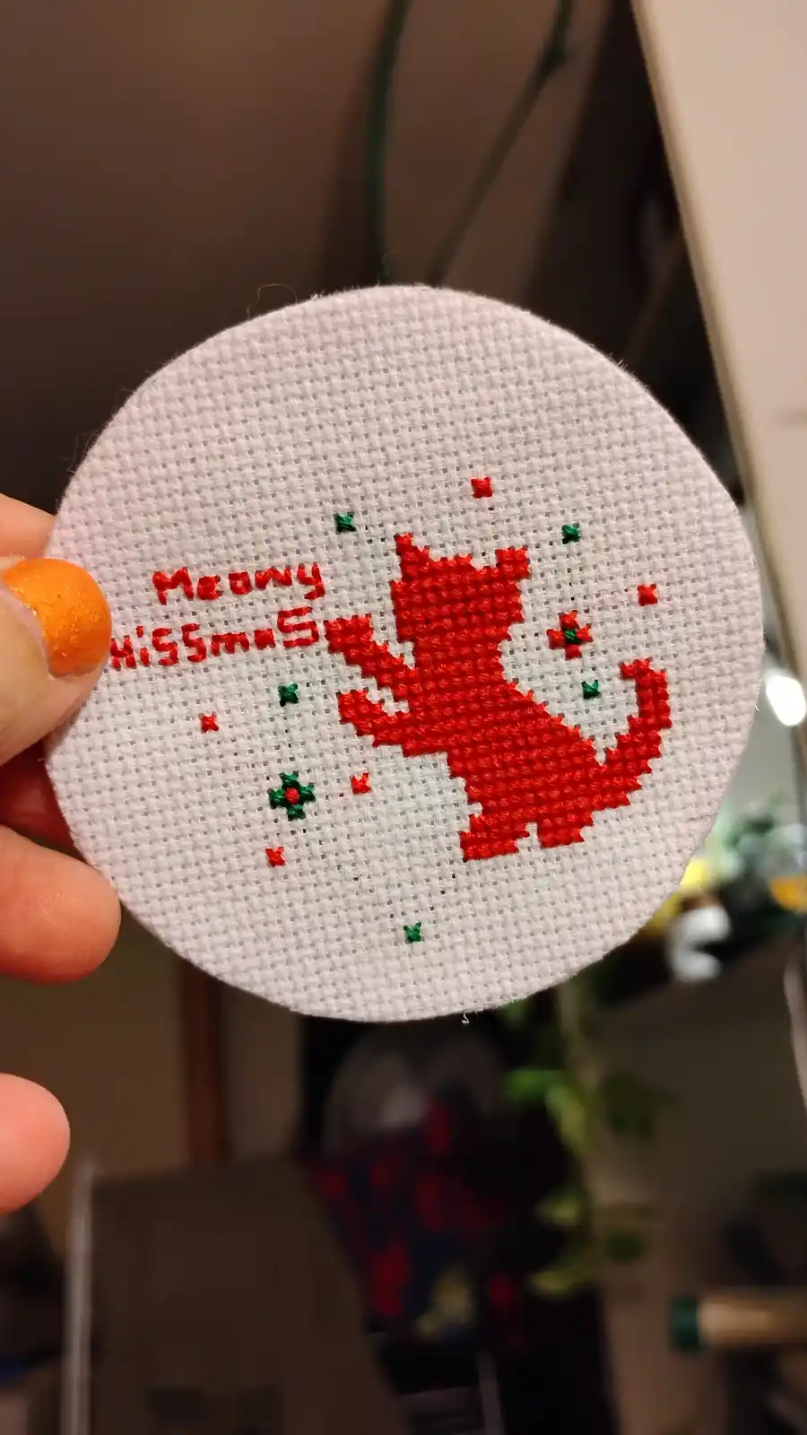 A hand holds a circular white cross-stitch piece featuring a red stitched cat silhouette and the text 'Meowy HiSSmas' in red…