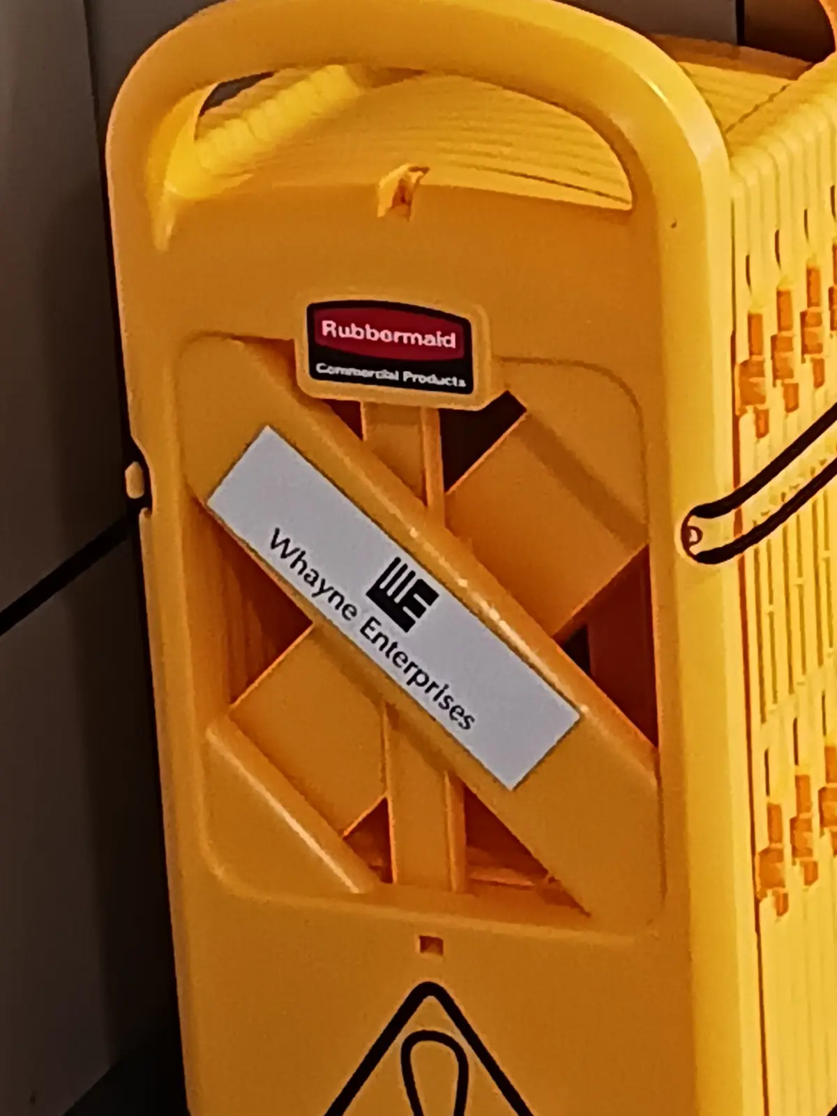 A close-up of a yellow Rubbermaid commercial mop bucket with a white diagonal sticker that reads 'Whayne Enterprises' and a…