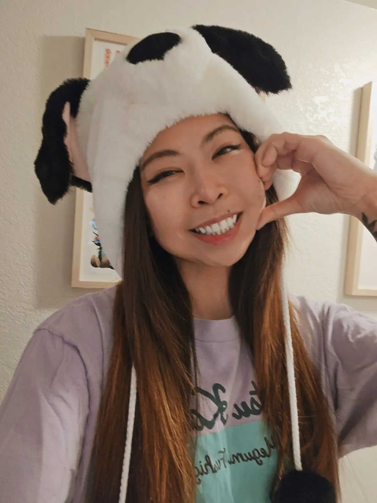 A person with long brown hair smiles while wearing a fluffy white and black panda-themed hat with ear flaps, making a…