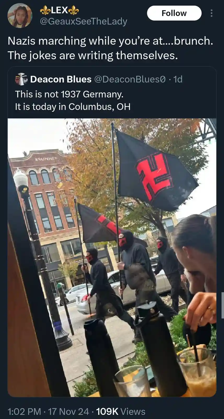 A photo taken from inside a cafe shows people in black clothing and masks holding black flags with red swastikas, marching…