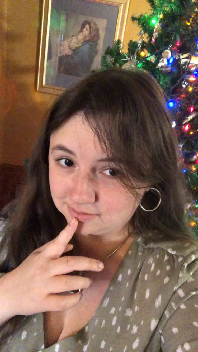 A young woman with dark hair and hoop earrings poses in front of a lit Christmas tree, her finger on her lips in a…