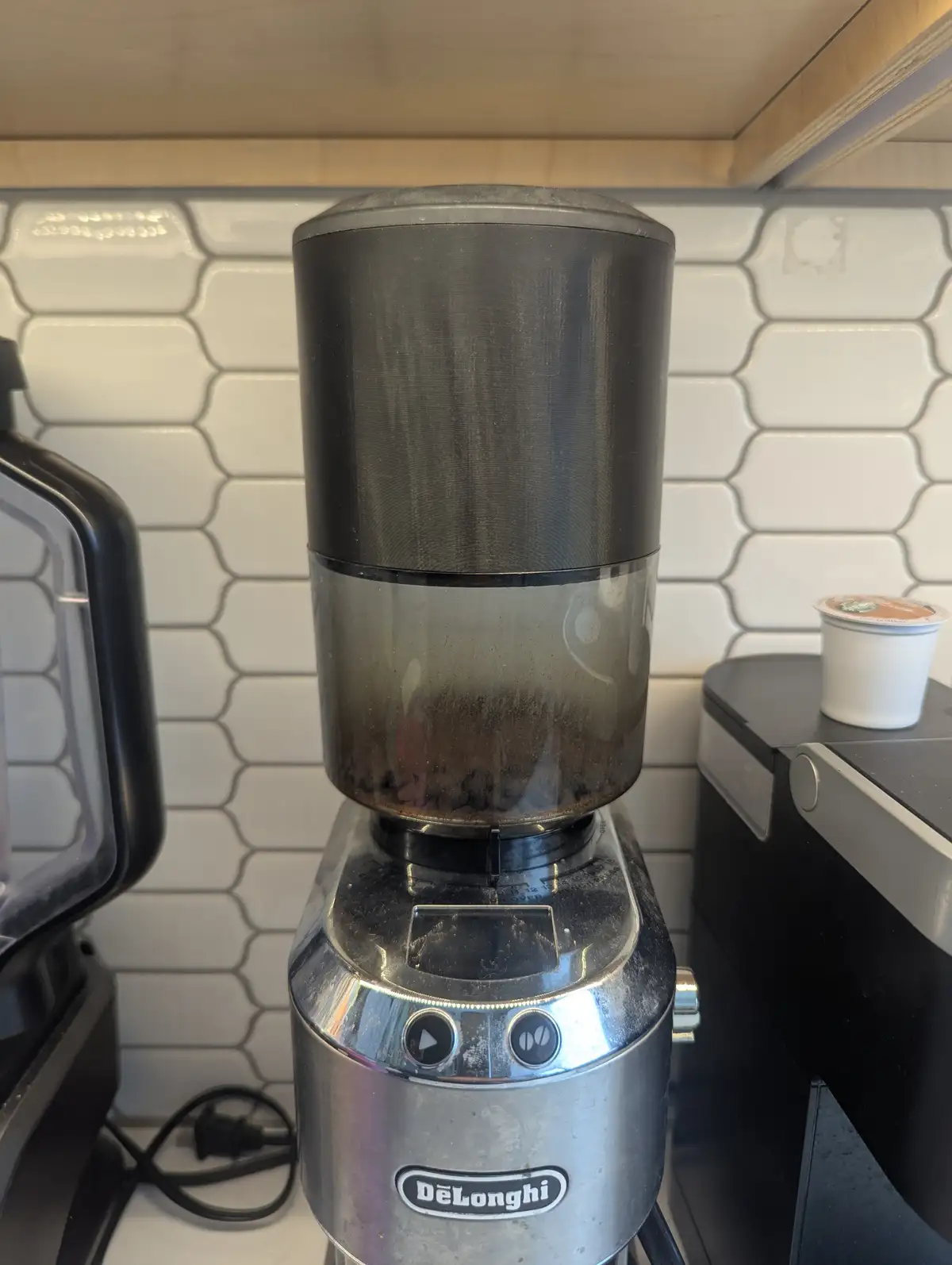 A DeLonghi coffee grinder sits on a kitchen counter with a white tiled backsplash, its transparent hopper filled with dark…