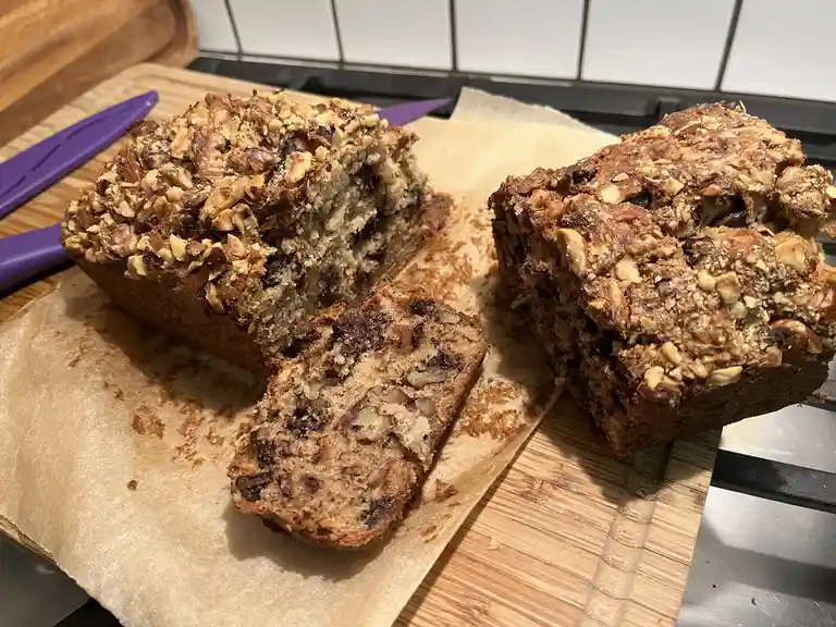 A close-up photo shows two loaves of banana bread with chopped nuts and dark chunks, one sliced, resting on parchment paper…