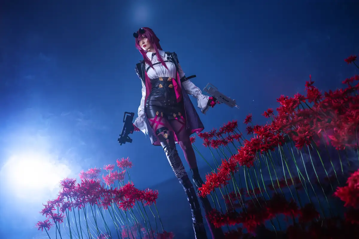 A cosplayer with pink hair and black-and-white outfit stands in a field of red spider lilies under a blue sky, holding two…