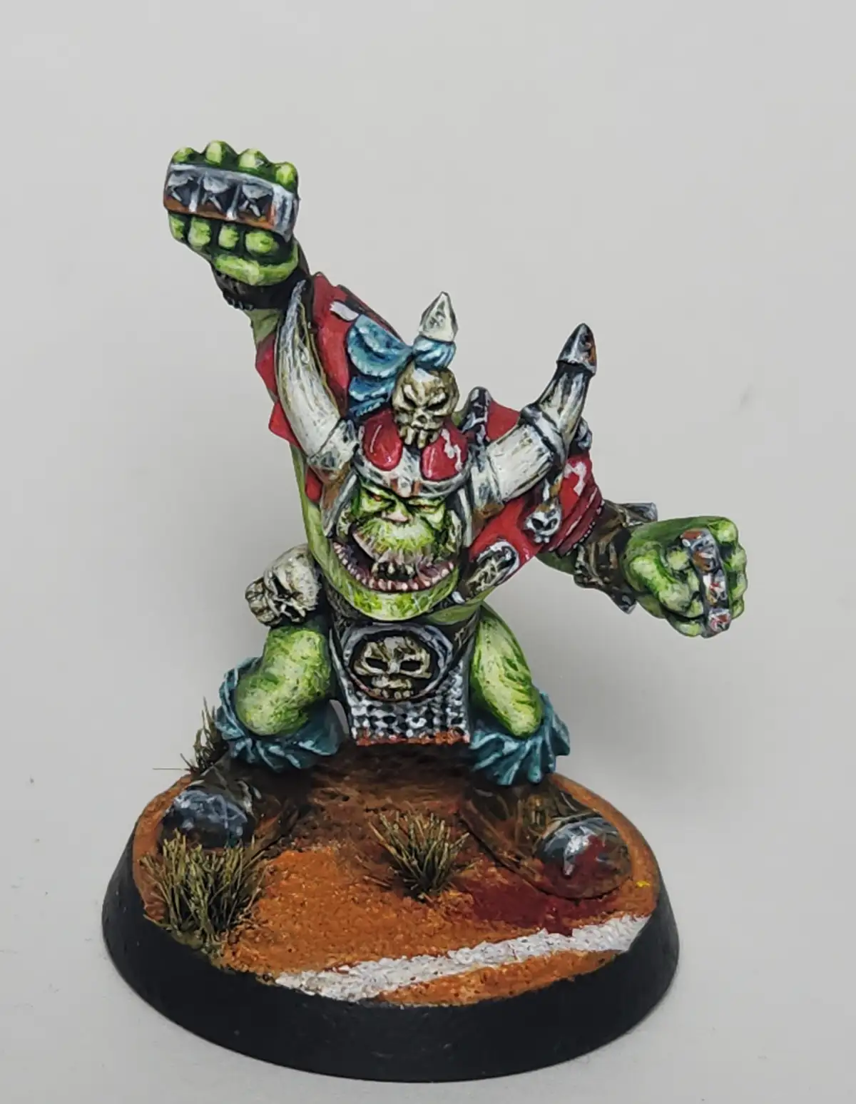 A painted green orc mini-figure with a skull helmet and red armor is posed aggressively, one fist raised, standing on a…