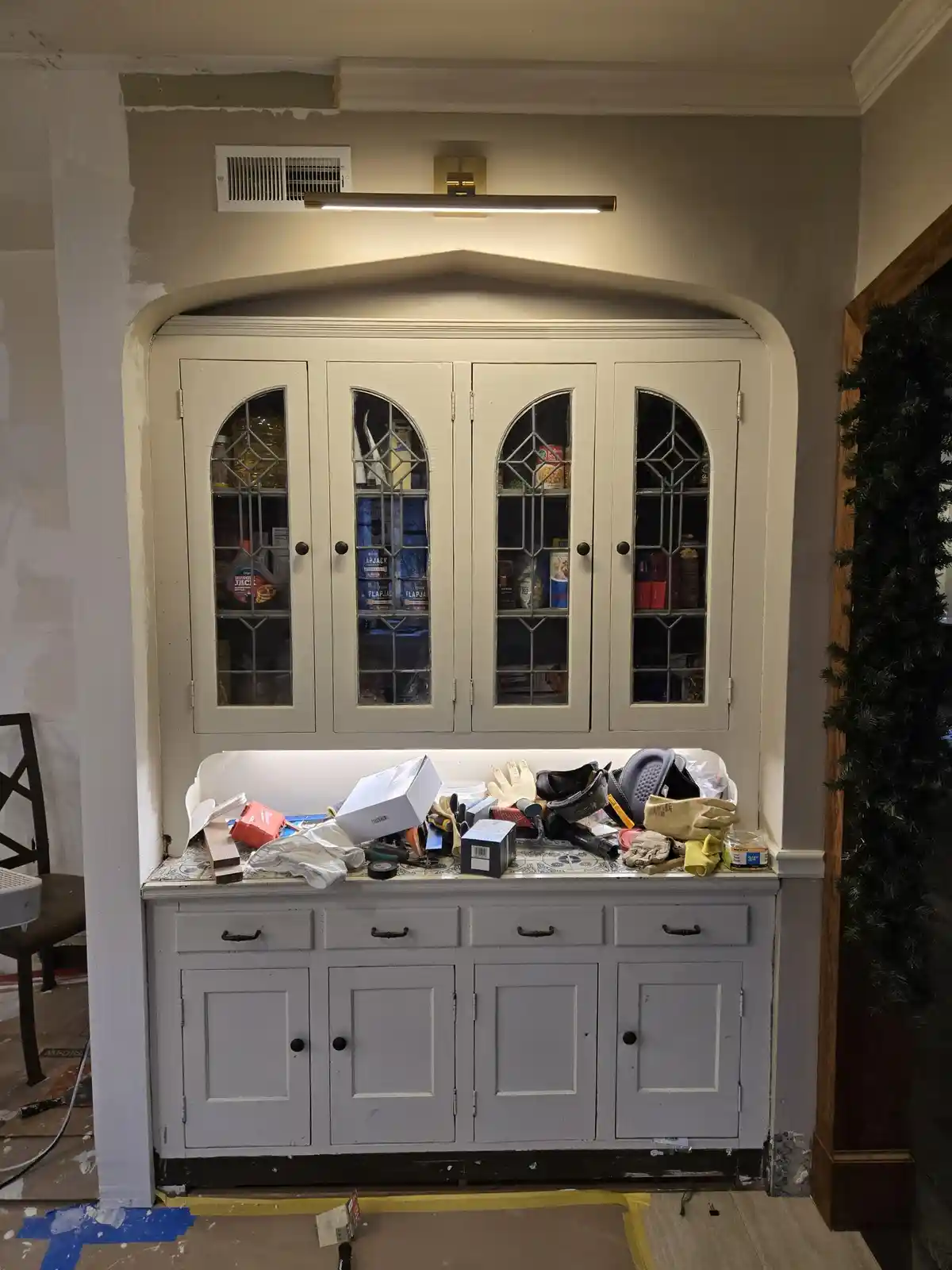 A white arched cabinet with glass doors filled with food items sits above a gray cabinet base cluttered with tools, gloves…