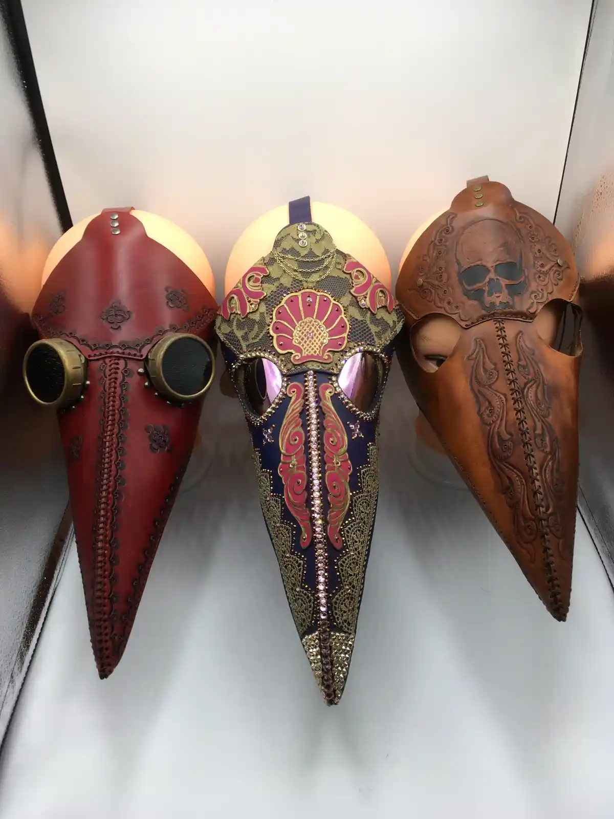 Three ornate, handcrafted plague doctor masks are displayed on mannequin heads against a white background. The left mask is…