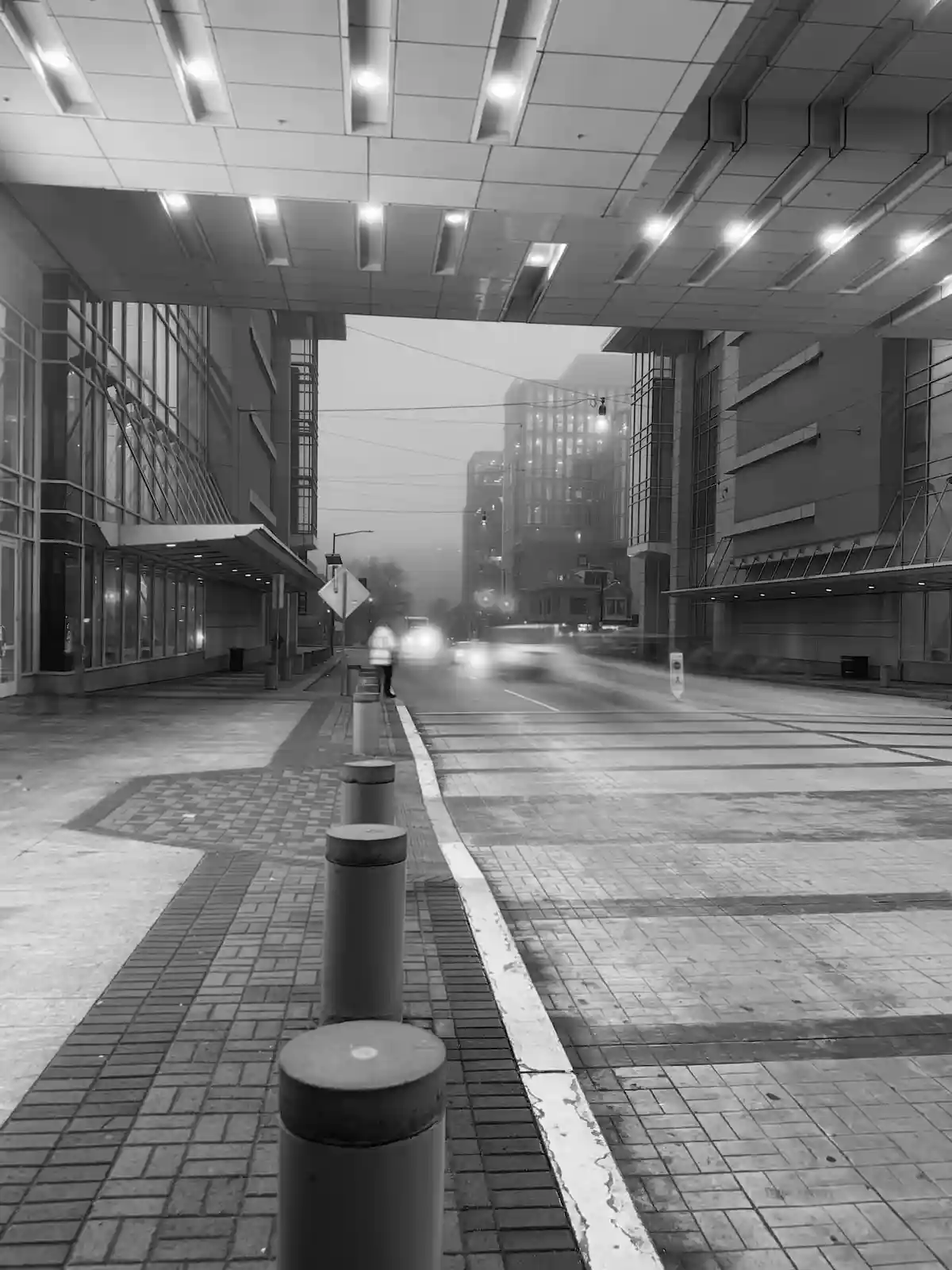A black-and-white, long-exposure photograph of a city street under a covered walkway, showing blurred motion of cars and a…