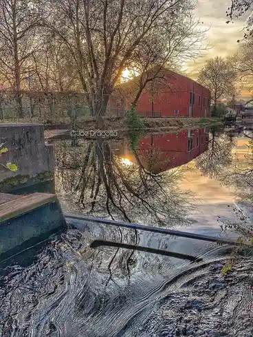 A serene photograph of a red barn and bare trees reflected in calm water at sunrise, with a small waterfall in the…