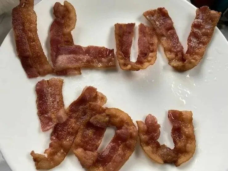 Crispy cooked bacon strips are arranged on a white plate to spell out the phrase 'i luv you' in lowercase letters. The bacon…