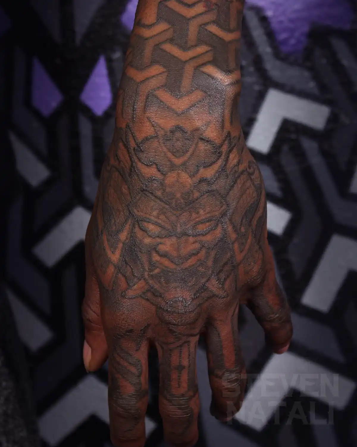 A close-up photo shows a person's hand and forearm covered in intricate black and brown tattoos, featuring a large demonic…
