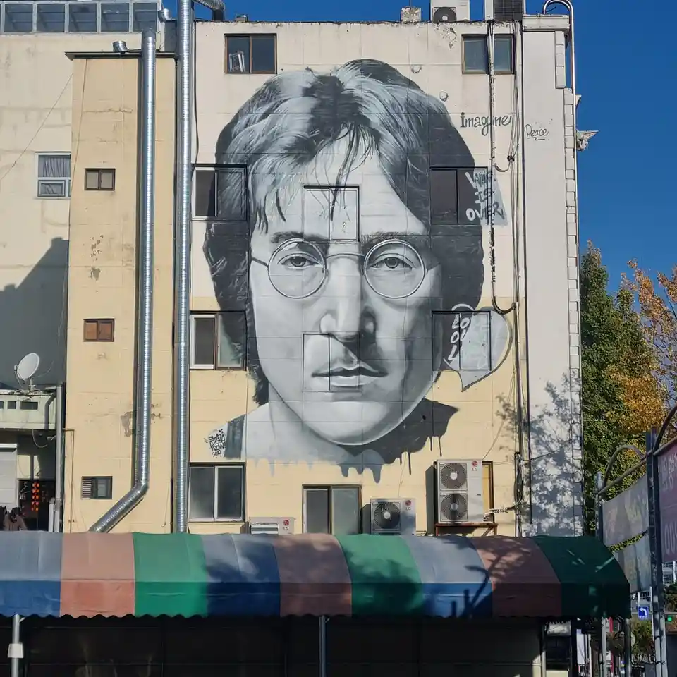 A large black-and-white mural of John Lennon's face is painted on the side of a building, with windows cutting through his…