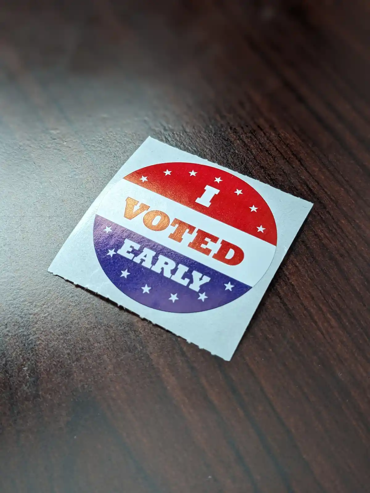 A close-up photo shows a circular 'I VOTED EARLY' sticker with red, white, and blue sections and white stars, resting on a…
