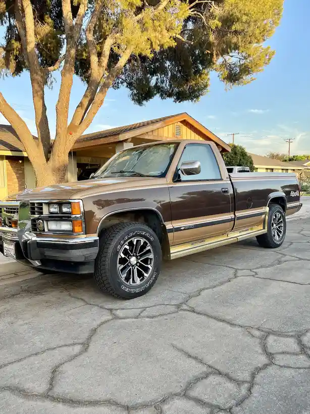 A brown and tan 1990s Chevrolet pickup truck is parked on a cracked driveway in front of a house with a large tree. The…