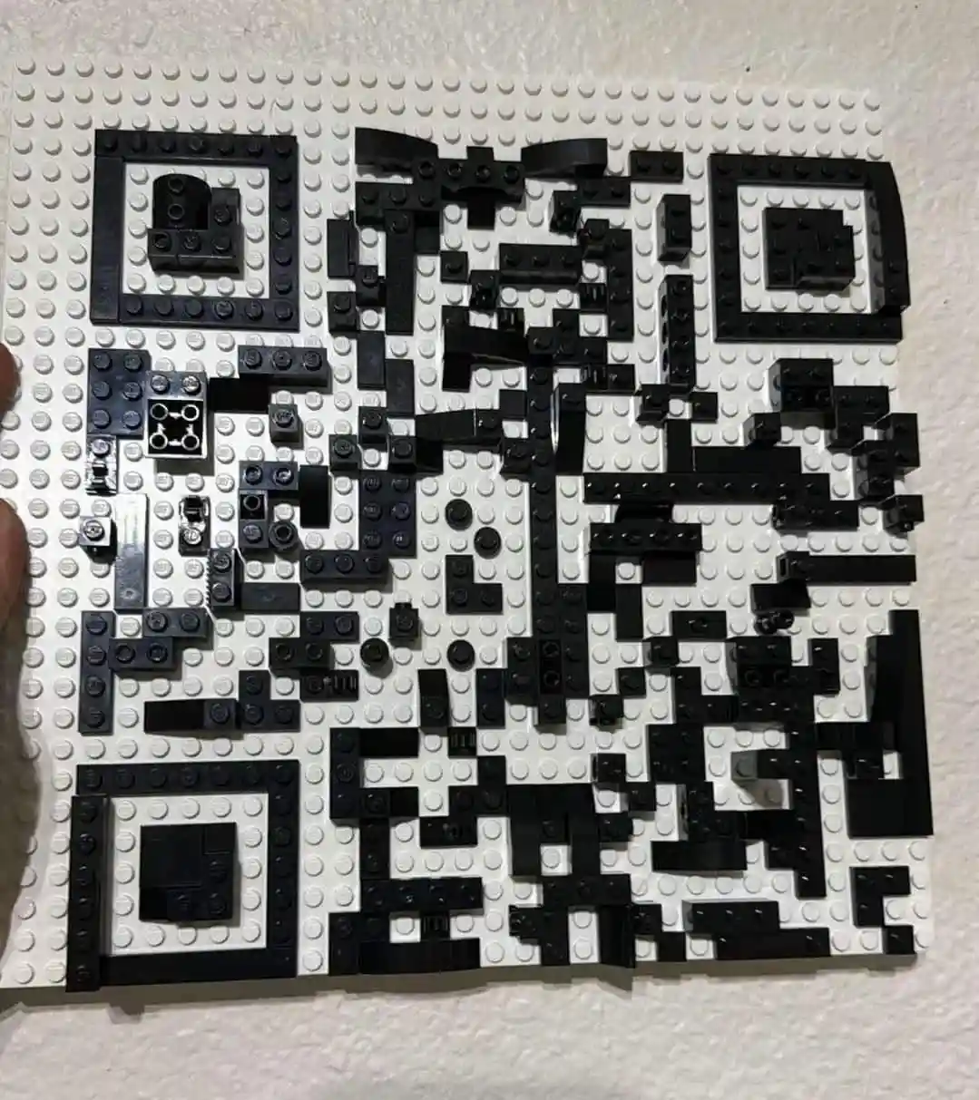 a qr code constructed from black and white lego bricks is displayed on a white lego baseplate, held by a person's thumb. the…