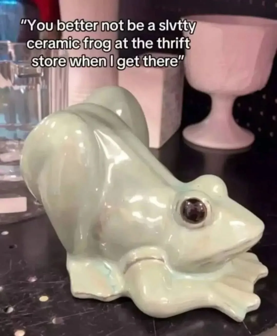 A glossy, light green ceramic frog figurine is shown sitting on a dark, speckled countertop, with various other decorative…
