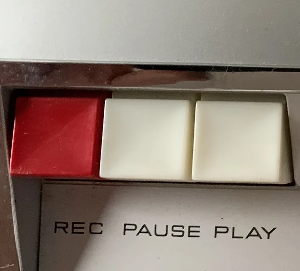 A close-up of a vintage tape recorder's control panel showing three buttons: a red 'REC' button on the left, and two white…