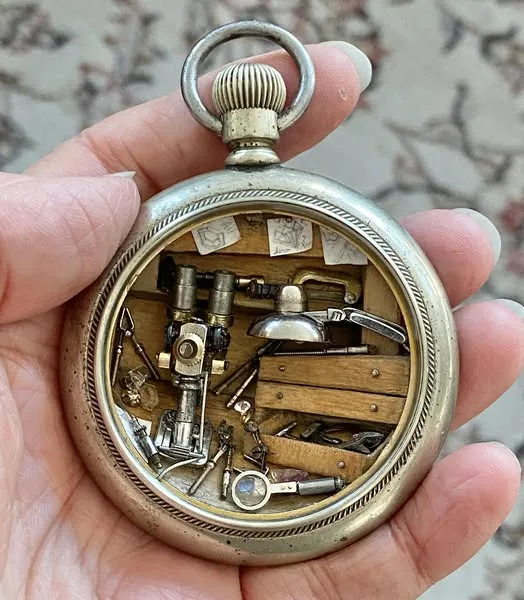 A hand holds an antique pocket watch that has been modified to contain a miniature workshop with tiny tools, wood pieces…
