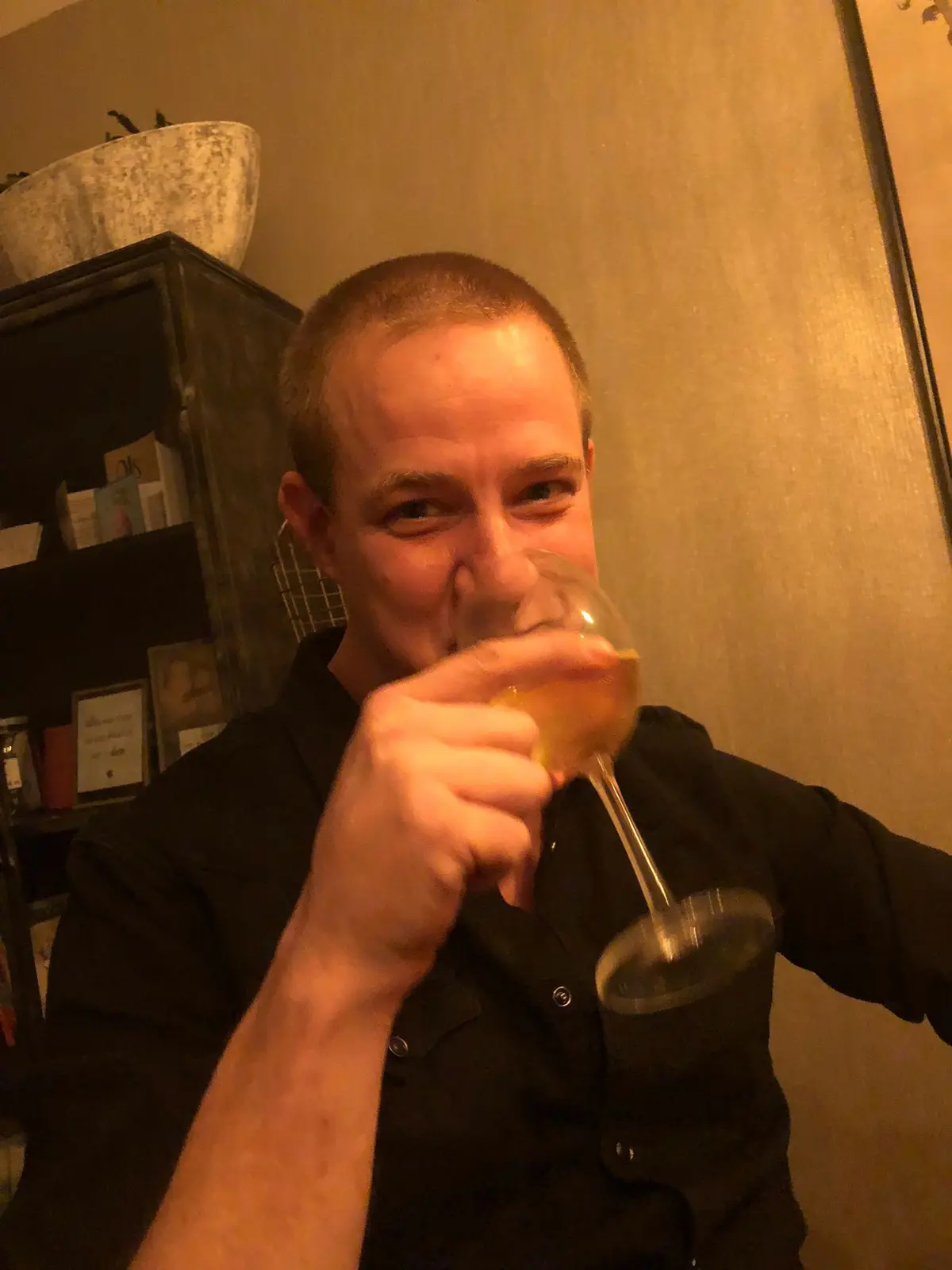 A man with short hair wearing a black shirt is holding a wine glass to his nose, appearing to smell the contents with a…